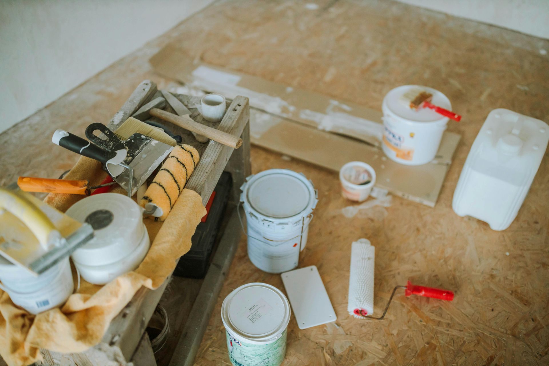 Painting supplies on wooden floor: paint cans, rollers, brushes, putty knives, and drop cloths.