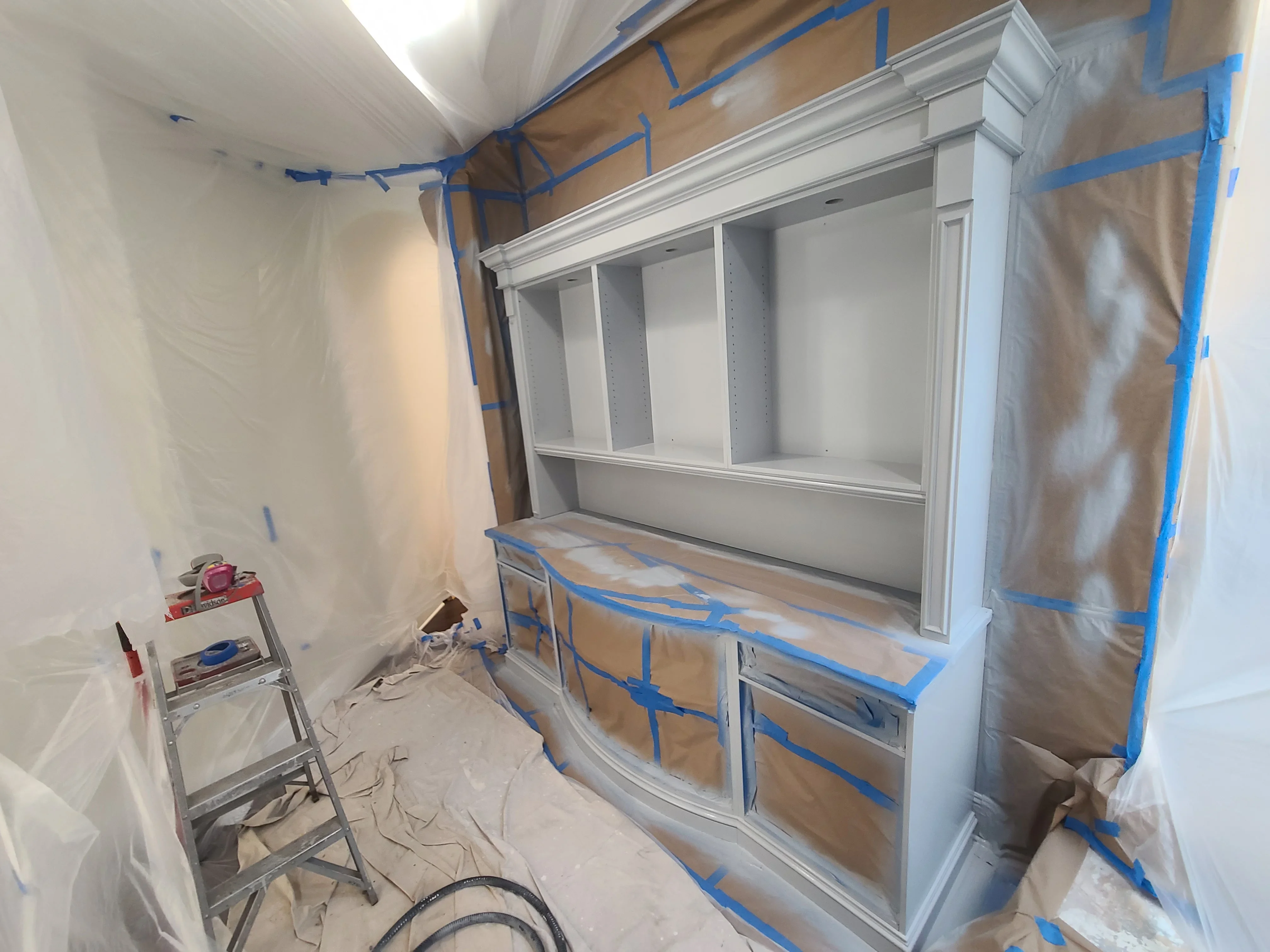 Interior room with primed drywall, blue painter's tape, kraft paper protection, and white built-in cabinet with shelving