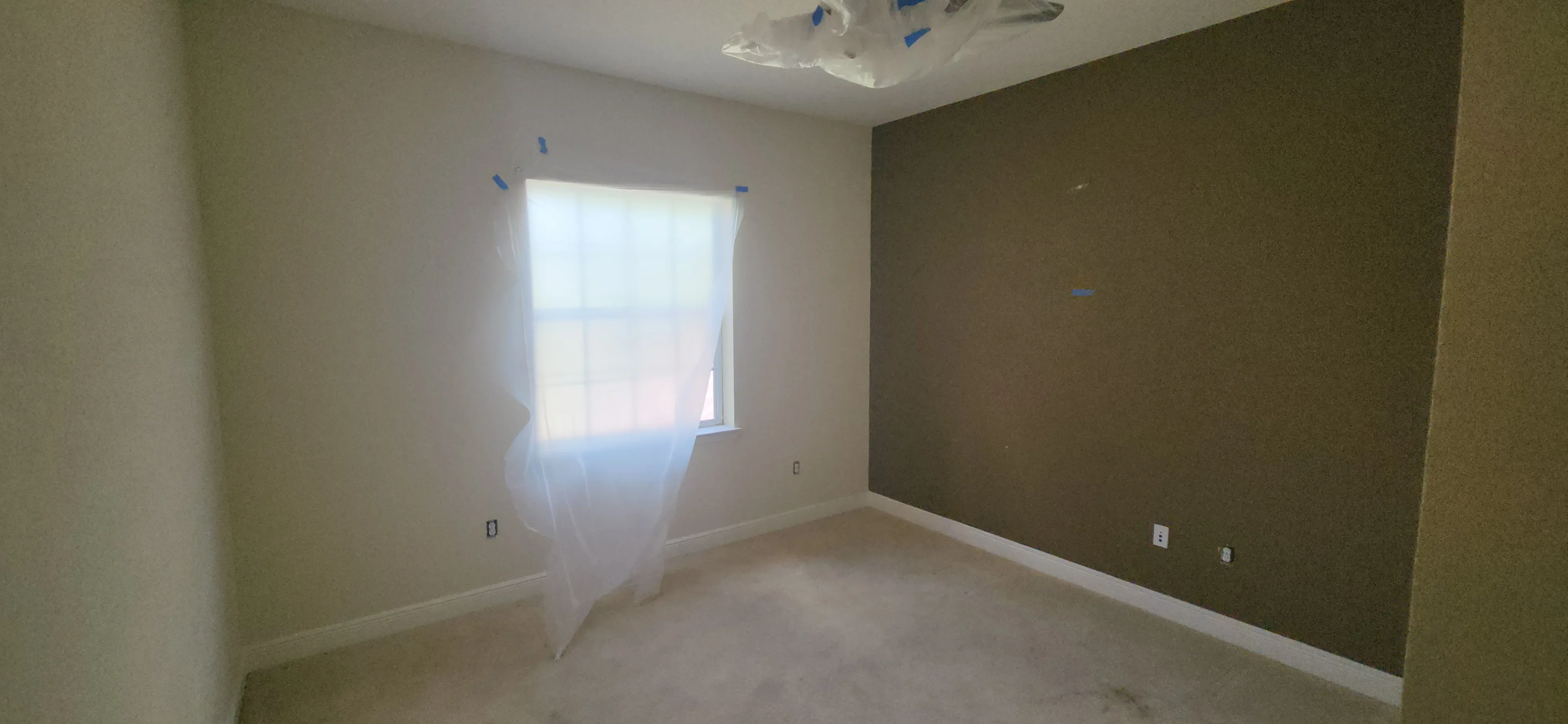 Bedroom interior with olive-green accent wall primer, protective plastic sheeting, and painter's tape on ceiling