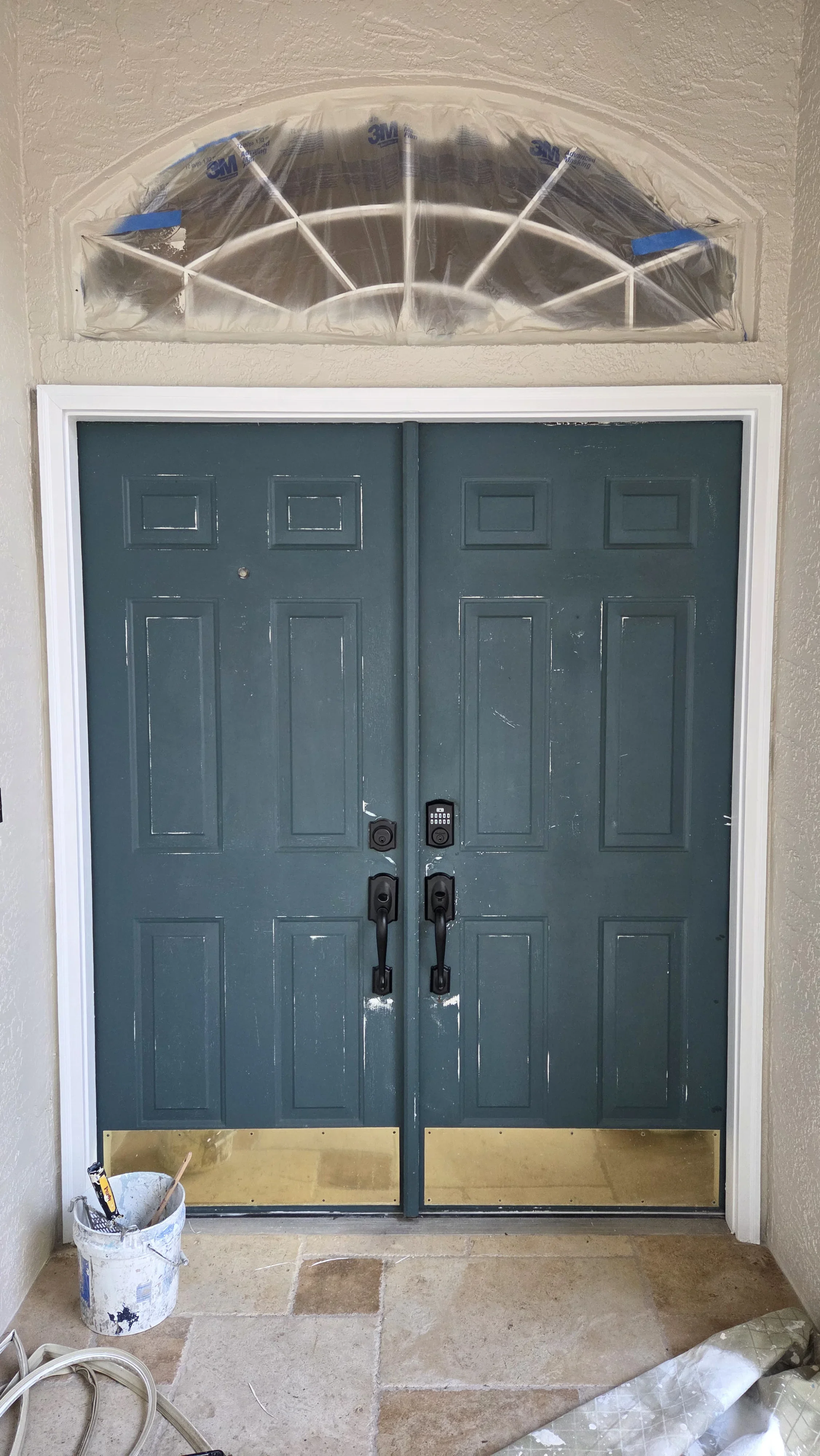 Double blue doors with six-panel design, transom window, brass kick plates, and protective plastic sheeting during painting preparation